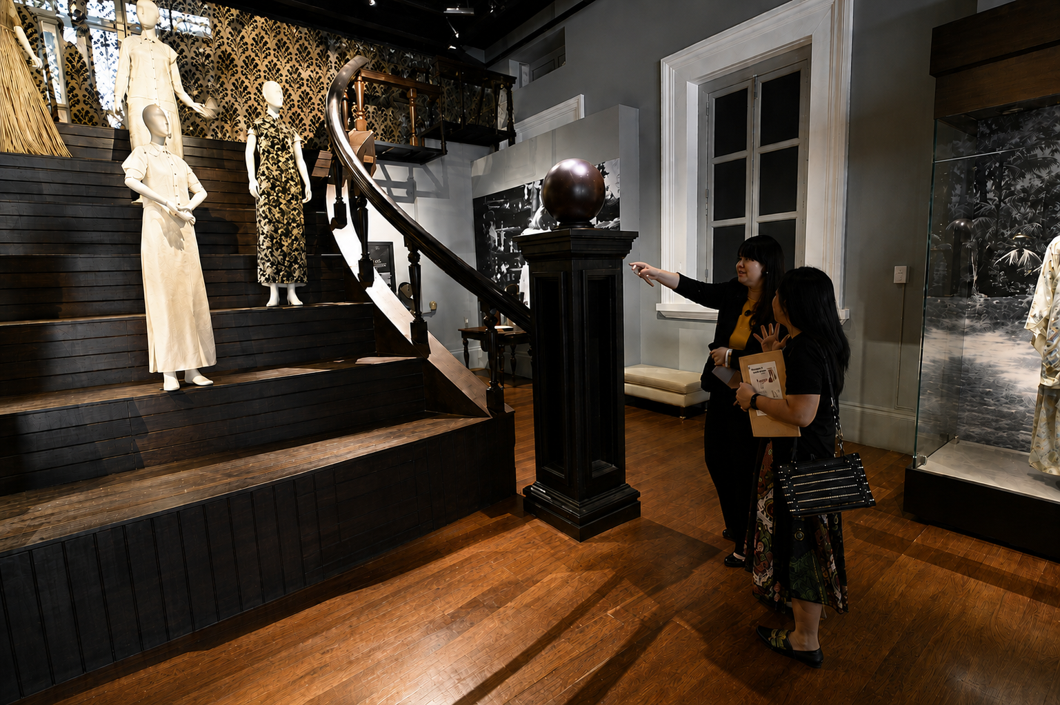 Two visitors examine a museum exhibit featuring a staircase display with historical mannequins wearing period costumes.
